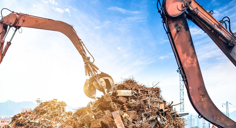 Excavators lifting and sorting scrap metal at a recycling facility under a clear blue sky.